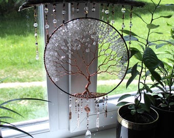 May include: A handcrafted wall hanging featuring a copper wire tree of life design within a black circular frame. Clear crystal beads and various other beads dangle from the frame and a natural wood branch. The artwork is displayed in front of a window.
