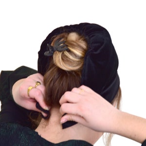 May include: A woman is putting on a black velvet hair covering. The hair covering is a circle shape and is being held in place with a black hair clip.