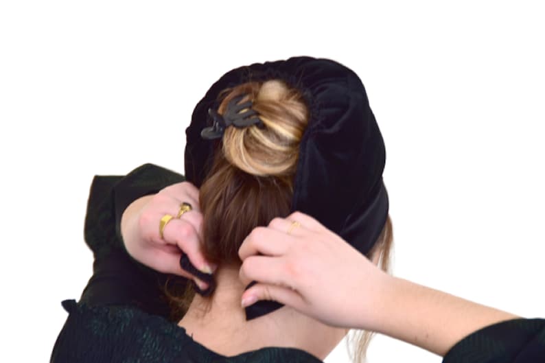 May include: A woman is putting on a black velvet hair covering. The hair covering is a circle shape and is being held in place with a black hair clip.