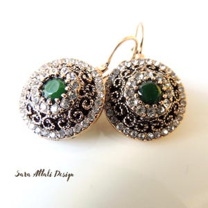 May include: A pair of gold-toned earrings with a central green stone surrounded by clear crystals. The earrings have a circular design with intricate filigree detailing. The text "Sara Attali Design" is visible.
