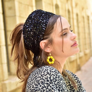 May include: A black sequined headband with a gathered design. The headband is worn by a person with long brown hair and yellow beaded earrings.