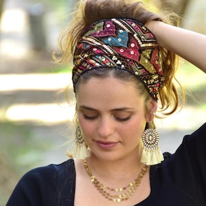 May include: A colourful headband with sequins and embroidery. The headband is black with a variety of colours including pink, blue, gold, and red. The headband is worn by a person with brown hair.