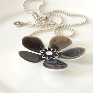 以下が含まれることがあります： 銀色のチェーンネックレスに黒い花のペンダントが付いている。花は5枚の花びらがあり、中央に小さなラインストーンが埋め込まれている。