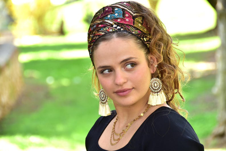 May include: A woman wearing a black top with a colourful patterned headband and large tassel earrings. The headband has sequins and embroidery.