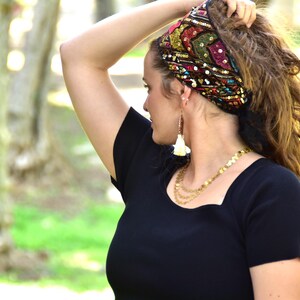 May include: A woman with curly brown hair is wearing a black top with a square neckline. She is wearing a colourful headband with sequins and a pair of white tassel earrings. She is also wearing a gold chain necklace.