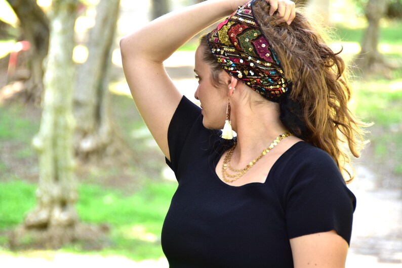 May include: A woman with curly brown hair is wearing a black top with a square neckline. She is wearing a colourful headband with sequins and a pair of white tassel earrings. She is also wearing a gold chain necklace.