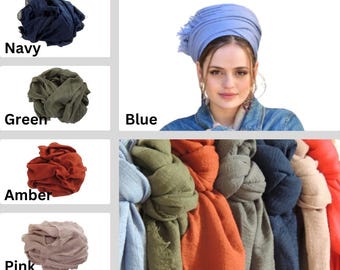 Op de afbeelding: Een verscheidenheid aan kleurrijke hoofddoeken en sjaals. Kleuren zijn onder meer marineblauw, groen, amber en roze. Een vrouw draagt een blauwe hoofddoek. De afbeelding toont ook een verzameling sjaals in verschillende kleuren.