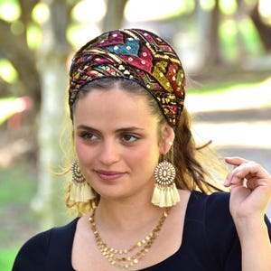 May include: A woman wearing a black headband with colourful embroidery and gold sequins. She is also wearing a black top and cream tassel earrings.