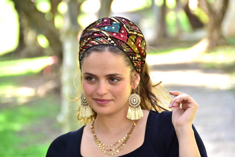 May include: A woman wearing a black headband with colourful embroidery and gold sequins. She is also wearing a black top and cream tassel earrings.