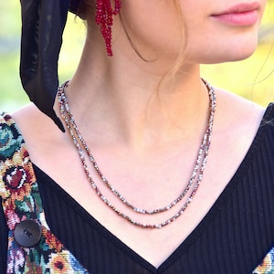 May include: A close-up of a woman wearing a black top with a floral pattern on the straps. She is wearing a double-strand necklace with small, iridescent beads.