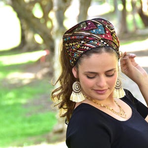 May include: A woman wearing a colourful head wrap with gold and silver embellishments. The head wrap is made of fabric and has a black base. The woman is wearing a black shirt and has long brown hair.