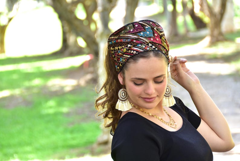 May include: A woman wearing a colourful head wrap with gold and silver embellishments. The head wrap is made of fabric and has a black base. The woman is wearing a black shirt and has long brown hair.