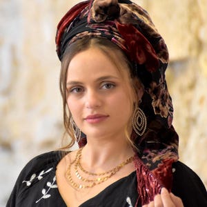 Pode incluir: Uma mulher usando um top preto com bordados florais brancos e um colar de ouro em várias camadas. Ela também está usando um véu com um padrão floral em tons de bordô, marrom e preto, e brincos pendurados.