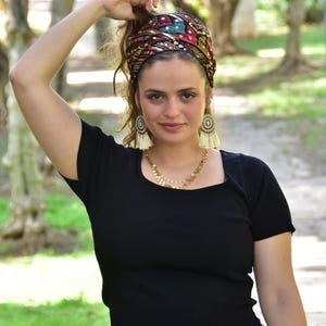 May include: A woman wearing a black top with a colourful patterned headband. The headband has sequins and is tied in a knot at the top of her head. She is wearing gold earrings with white tassels.