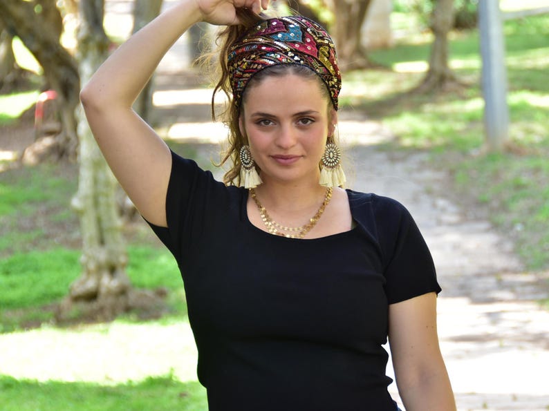 May include: A woman wearing a black top with a colourful patterned headband. The headband has sequins and is tied in a knot at the top of her head. She is wearing gold earrings with white tassels.