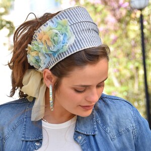Puede incluir: Una diadema a rayas azules y blancas con un gran adorno floral azul, verde y amarillo. La diadema la lleva una persona con el pelo castaño.