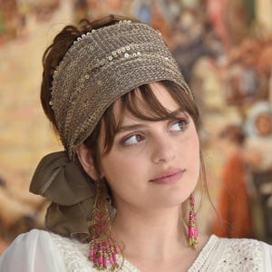 May include: A woman wearing a brown headband with sequins and a long pair of pink and gold earrings. She is wearing a white top with a lace pattern.