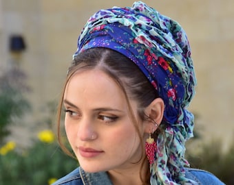 May include: A blue floral headscarf with a ruffled edge. The headscarf is wrapped around the head and tied at the back. The scarf has a floral pattern in shades of red, pink, and green. The person is wearing a denim jacket and red beaded earrings.