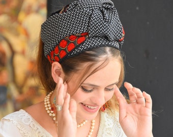 May include: A woman wearing a black and white patterned head scarf with red accents. She is wearing a white lace top and a pearl necklace.
