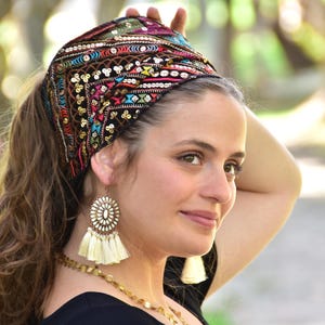 May include: A woman wearing a black headband with colourful embroidery and sequins. The headband is tied in a knot at the top of her head. She is also wearing large, white tassel earrings.