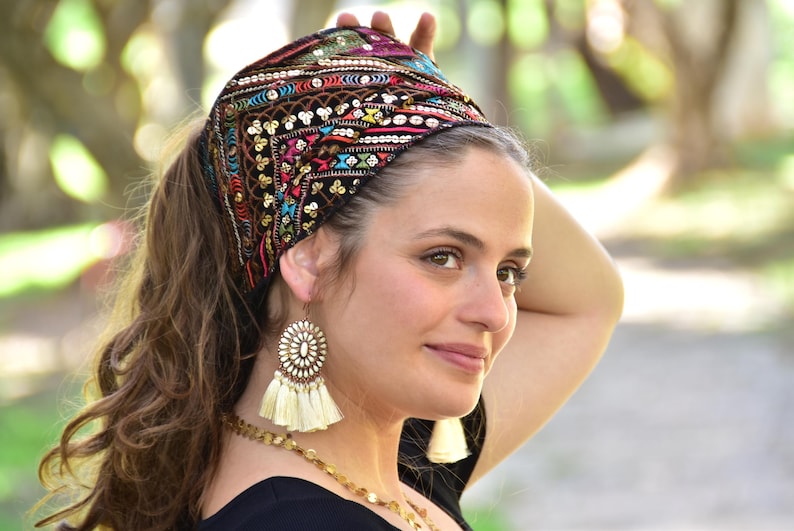 May include: A woman wearing a black headband with colourful embroidery and sequins. The headband is tied in a knot at the top of her head. She is also wearing large, white tassel earrings.