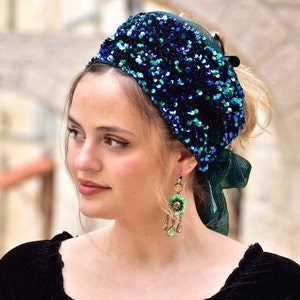 Puede incluir: Una mujer lleva una diadema de lentejuelas azules y verdes con un lazo de tela verde. También lleva pendientes largos colgantes con detalles verdes y dorados.