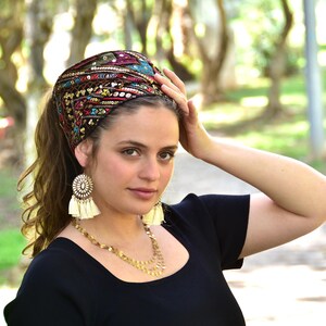 May include: A woman wearing a black top with a colourful embroidered head scarf with sequins. She is wearing gold earrings with white tassels.