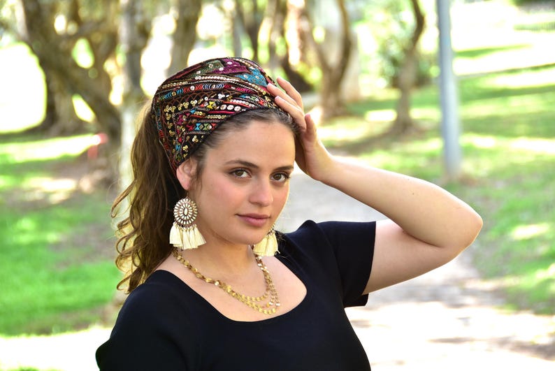 May include: A woman wearing a black top with a colourful embroidered head scarf with sequins. She is wearing gold earrings with white tassels.
