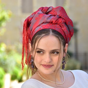 May include: A woman wearing a red and gray patterned headscarf with long red tassels. She is wearing a white shirt and a pearl necklace.