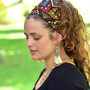 May include: A woman with long brown curly hair wearing a black top and a colourful beaded headband. She is wearing large white tassel earrings.
