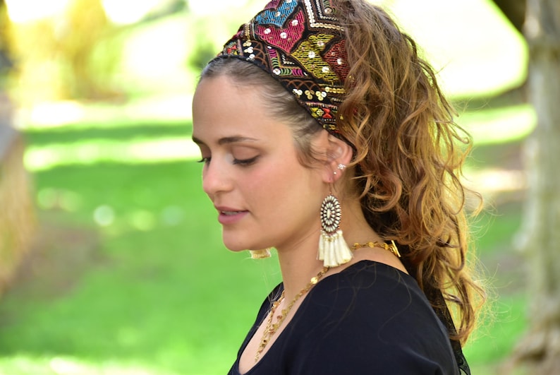 May include: A woman with long brown curly hair wearing a black top and a colourful beaded headband. She is wearing large white tassel earrings.
