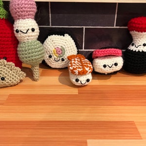 May include: A collection of crocheted amigurumi food toys, including a red hot sauce bottle, a green dango, a white and pink sushi roll, an orange and white sushi roll, a pink and white sushi roll, and a black soy sauce bottle.