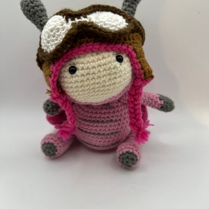 May include: A pink crocheted toy with a brown and white aviator hat. The toy has gray crocheted feet and pink crocheted hair.