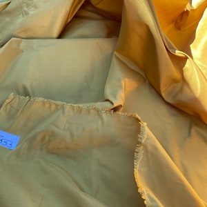 Puede incluir: Primer plano de un gran trozo de tela de color amarillo dorado con borde festoneado. La tela está doblada y drapeada, creando pliegues y arrugas suaves. La tela es lisa y sedosa, con un ligero brillo.