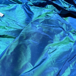 May include: Close-up of iridescent fabric in shades of blue and green. The fabric has a shimmering, silky appearance with visible folds and creases, creating a textured effect. The material reflects light, showcasing a color-shifting quality.