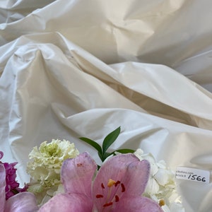 May include: A close-up of a bouquet featuring a large, light pink lily with visible stamens, surrounded by white and green flowers. The background is a cream-colored, textured fabric. A tag with the text "Silks4less.com Article # 1566" is visible.