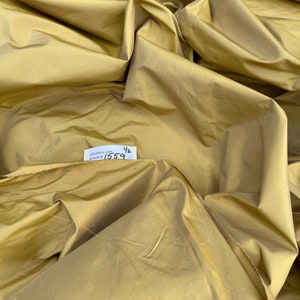 Puede incluir: Un primer plano de una tela de seda de color amarillo dorado. La tela está doblada y drapeada, mostrando su textura y brillo. Se puede ver una pequeña etiqueta blanca con el texto "silkdress.com Article # 1559 1/2".