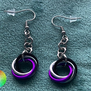 May include: A pair of silver earrings with a black, silver, and purple interwoven ring design. The earrings are hanging from a silver chain.