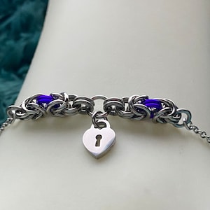 May include: A silver chain bracelet with a heart-shaped charm featuring a keyhole. The bracelet has two purple beads on each side of the charm.