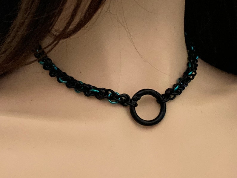 以下が含まれることがあります： 黒とターコイズブルーのチェーンチョーカーに、大きな黒い金属製のリングが付いている。