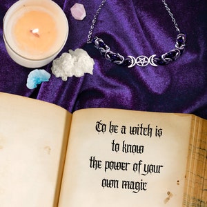 May include: An open book with the text "To be a witch is to know the power of your own magic" on a purple velvet background. There is a silver necklace with a triple moon and pentacle charm, a lit candle, and crystals on the surface.
