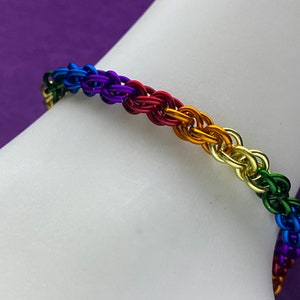 May include: A rainbow colored chainmail bracelet with a woven pattern. The bracelet is made of metal and is worn on a person's wrist.