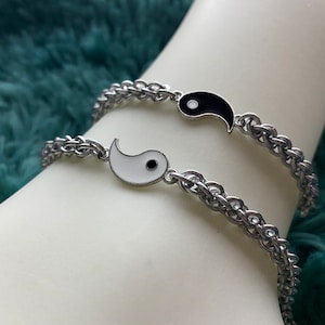 Puede incluir: Dos pulseras de cadena plateadas con dijes yin yang. Un dije es negro con un punto blanco, y el otro es blanco con un punto negro.