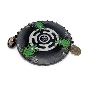 Puede incluir: Un objeto decorativo redondo y negro con un diseño de laberinto blanco y tres ranas verdes. Tiene una cadena de cuentas y un pequeño colgante ovalado. Texto: "In the shadows. Your wisdom is..."