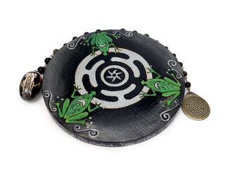 Puede incluir: Un objeto decorativo redondo y negro con un diseño de laberinto blanco y tres ranas verdes. Tiene una cadena de cuentas y un pequeño colgante ovalado. Texto: "In the shadows. Your wisdom is..."