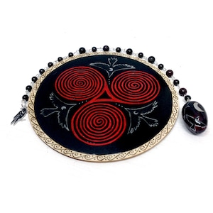 Peut inclure: Décoration murale ovale noire avec un motif de spirales rouges et une bordure décorative. Le motif comprend trois spirales rouges et des illustrations d'oiseaux argentés. Une chaîne de perles avec un pendentif est suspendue sur le côté. Le pendentif est une pierre sombre avec un motif d'oiseau et de lune.