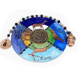 May include: A round, hand-painted decorative piece with a spiral center and deer imagery. The piece is divided into sections of blue, green, and yellow, with a beaded chain and acorn-shaped charm attached. The overall design has a rustic, artistic feel.