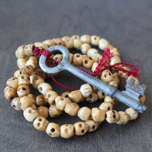 May include: A silver key with a skull-shaped bead necklace. The necklace is made of bone-like beads with a red string and tassel.