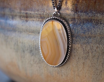 May include: A silver pendant necklace with a rope chain and an oval-shaped agate stone. The agate is a light brown color with white stripes.