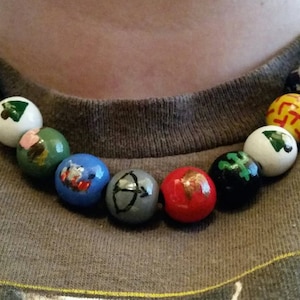 May include: A necklace made of wooden beads with different colored paint and designs. The beads feature a variety of symbols, including a tree, a shield, an arrow, and a house.